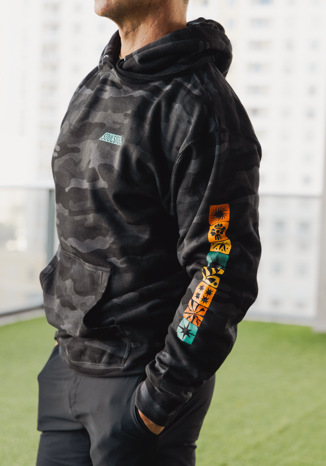 Image of athletic man with muscles in a gym wearing the Soulside Industry hoodie, a cozy hoodie with original artists design. The hoodie is black camo with colorful elements down the sleeve and on the back.