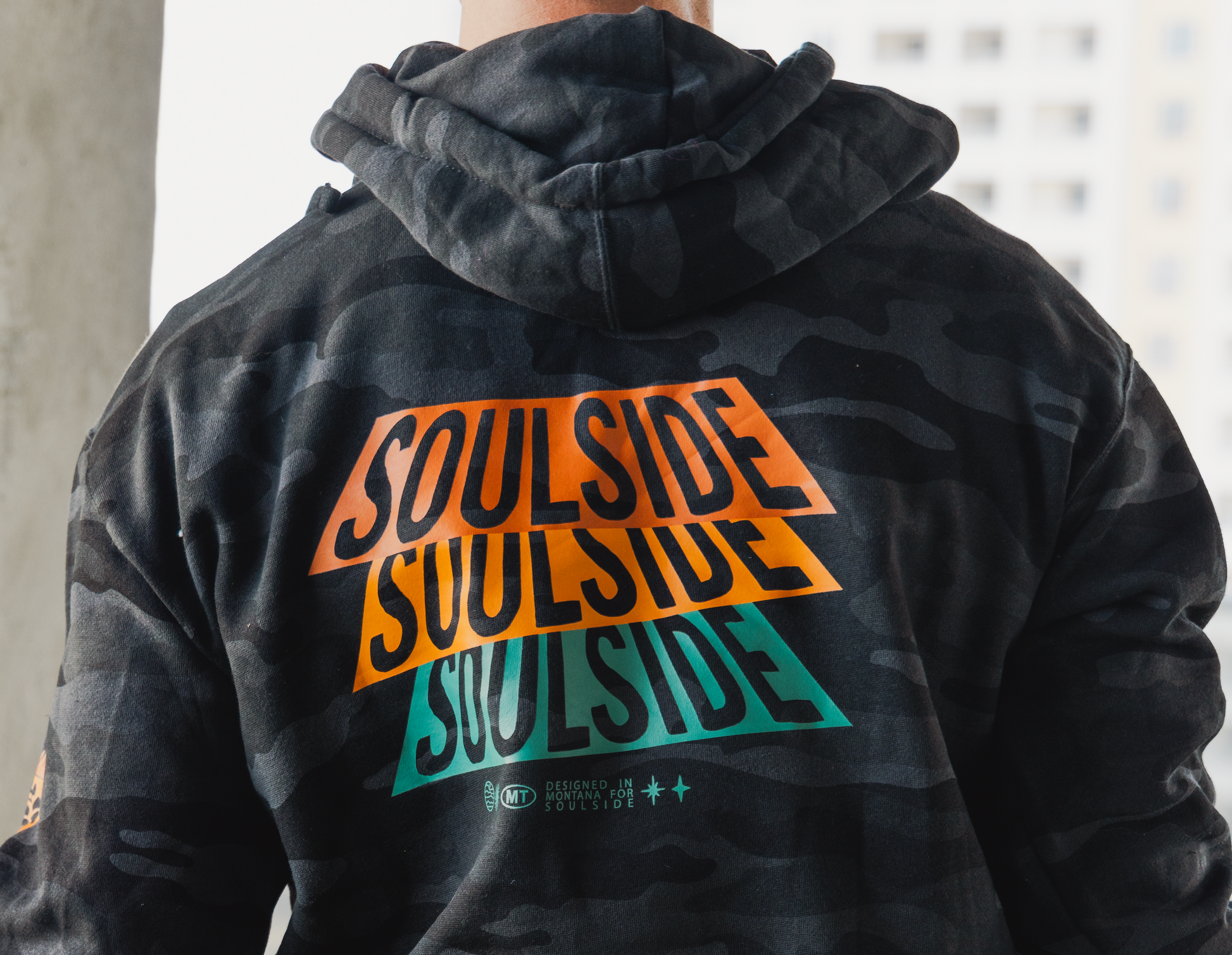 Image of athletic man in a gym wearing Soulside Industry hoodie. The image is of the back of the cozy black camo hoodie, showing the industry artist design on the back with orange, yellow and turquoise lettering that says Soulside.